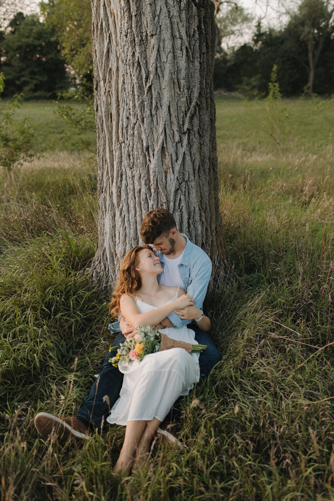 A couple sit under at tree during their engagement photo session at Pioneers Park in Lincoln, Nebraska. 