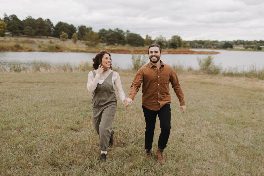 Man and woman holding hands and running away from the lake at Holmes Lake in Lincoln, Nebraska, during their engagement photo session. 