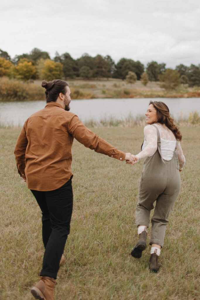 Man and woman holding hands and running to the lake at Holmes Lake in Lincoln, Nebraska, during their engagement photo session. 